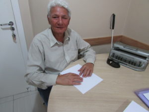 Descrição da Imagem: Um homem de cabelos grisalhos está sentado lendo um texto em braille apoiado sobre uma mesa redonda. Fim da descrição.