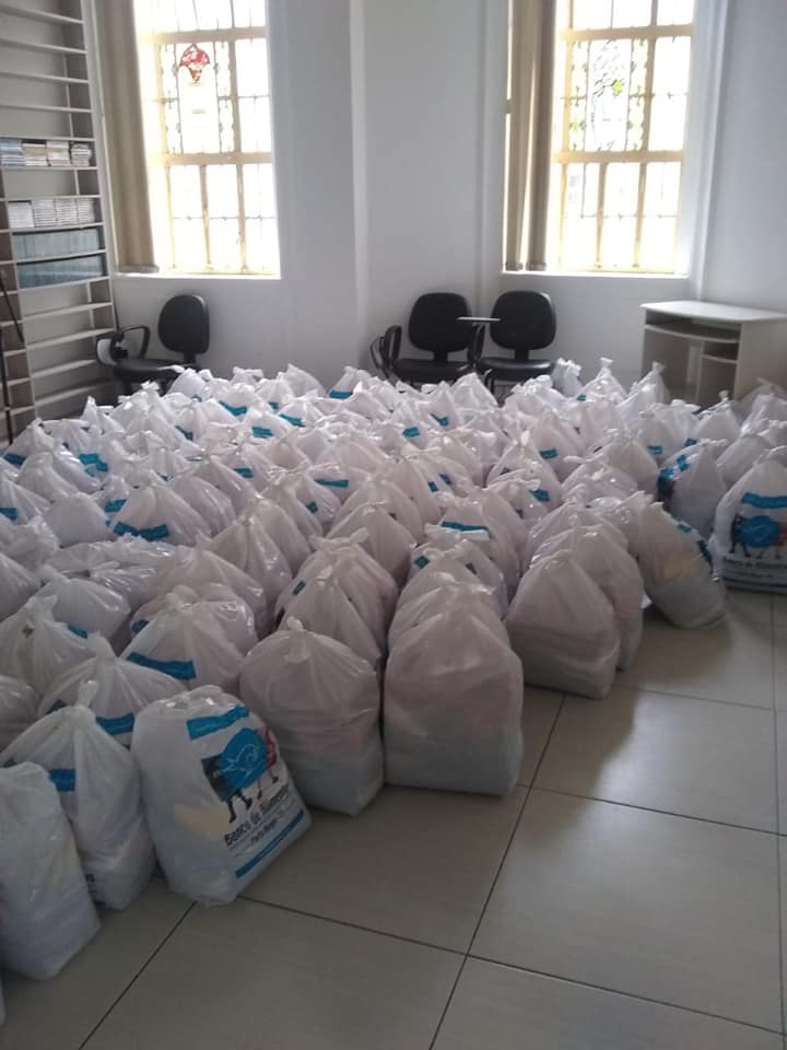 Descrição da imagem: foto de salão com dezenas de sacolas brancas de cestas básicas ao chão. Fim da descrição