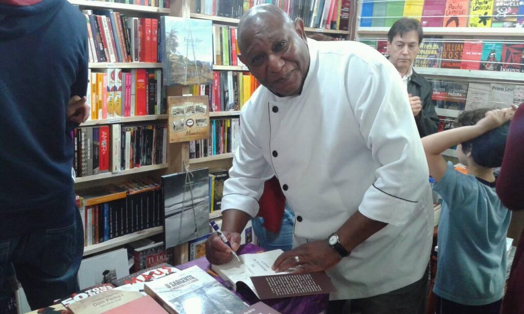 Foto do Chefe de cozinha Pedro Nascimento, autografando o seu livro de receitas. Ao fundo livros em uma prateleira. Fim da descrição