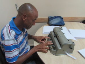Descrição da Imagem: Um homem sentado, ele está escrevendo em uma máquina Perkins, máquina de escrever em braille. Fim da descrição