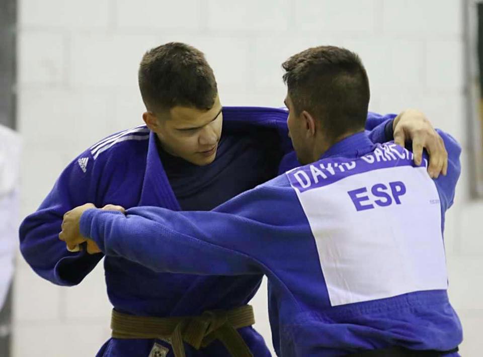 Descrição da imagem: Foto em orientação paisagem destaca Judoca Anderson Wassian em treino com o judoca David Garcia da Espanha. Os dois atletas estão em pé e usam kimonos azuis. Eles travam disputa de pegada. Fim da descrição.