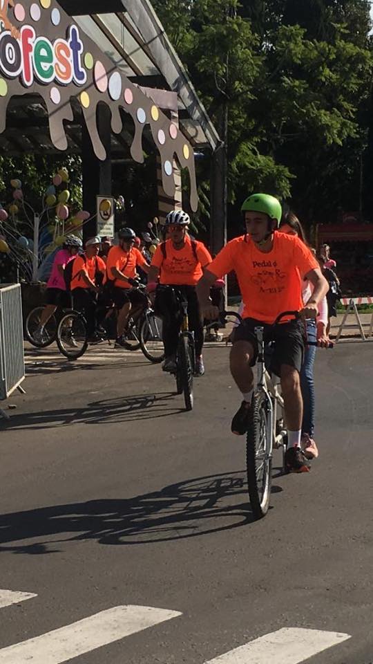 Descrição da imagem: Foto em orientação retrato destaca ciclistas de camisetas cor laranja com logo Pedal da ACERGS na altura do peito. Eles pedalam bicicletas duplas e ao fundo e no alto o pórtico da ChocoFest.
Fim da descrição.
