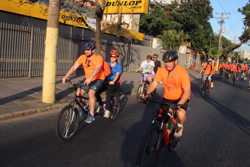 Descrição da imagem: foto orientação paisagem destaca grupo de ciclistas alinhado em uma faixa de via pública. Eles estão em bicicletas duplas e usam predominantemente camisetas laranja.
Fim da descrição