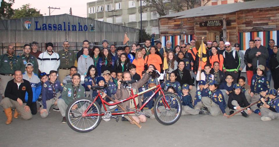 Descrição da imagem: foto em orientação paisagem destaca grupo de ciclistas e escoteiros posando para foto com uma bicicleta de dois lugares tanden a frente. Fim da descrição.
