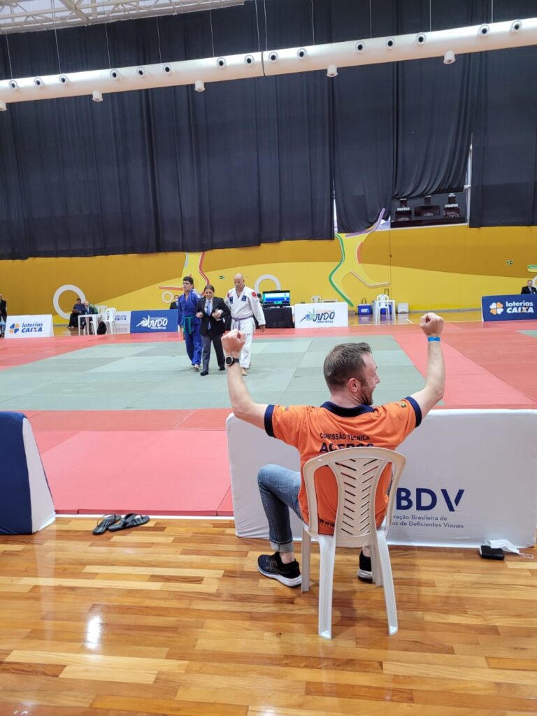 Descrição da imagem: Foto dos dois lutadores mais ao fundo da imagem sendo conduzidos para fora do tatame. Ao lado esquerdo, lutador de kimono azul e faixa verde, no centro a pessoa que os conduz e à direita, o atleta Sidinei de kimono branco e faixa roxa. Mais à frente na imagem, treinador sentado de costas com os braços para cima em um momento de comemoração. Fim da descrição.