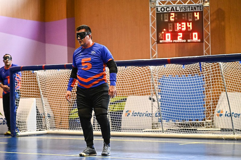 Alex Gaúcho está de pé na região central do seu lado da quadra. Ele veste camisa de manga longa azul e calça preta. O número 5, na altura do peito, está escrito em laranja.