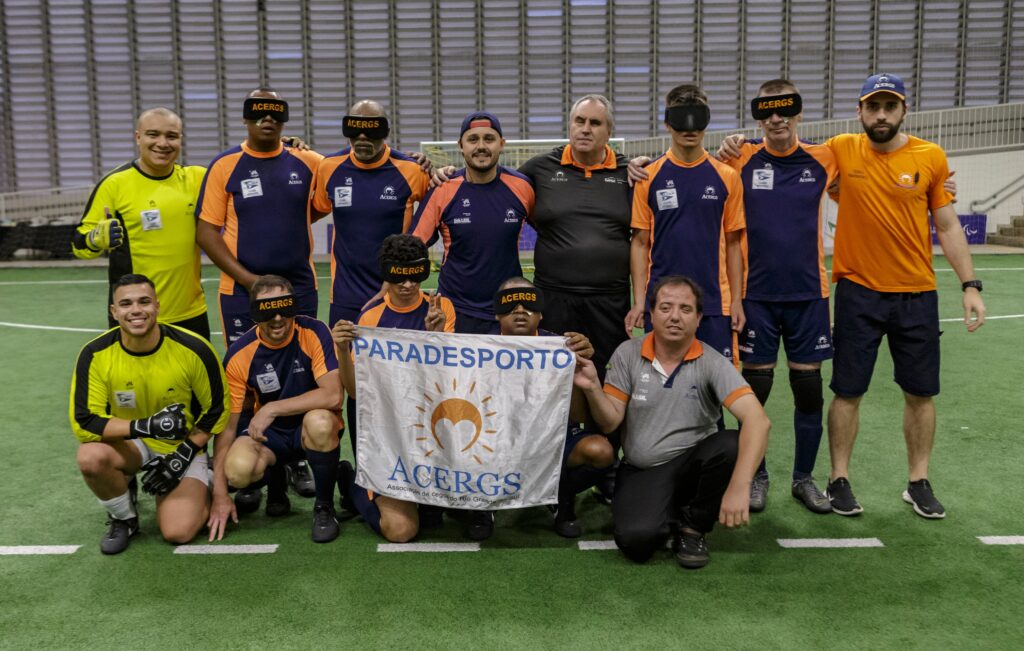 A imagem mostra um grupo de pessoas posando para uma foto em um campo de futebol coberto. Eles estão usando uniformes esportivos, predominantemente nas cores azul e laranja. Alguns dos integrantes estão usando vendas pretas nos olhos com a palavra "ACERGS" escrita em laranja. Na frente do grupo, duas pessoas estão segurando uma bandeira que diz "PARADESPORTO ACERGS" com um logotipo que parece um sol estilizado. A bandeira também tem a inscrição "Associação de Cegos do Rio Grande do Sul". Ao fundo, há uma estrutura metálica que parece ser parte de um ginásio.