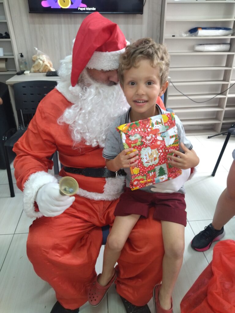 Na imagem, há um Papai Noel sentado em uma cadeira, segurando um sino dourado em uma das mãos. Ele está vestido com o tradicional traje vermelho e branco, incluindo um gorro vermelho. No colo do Papai Noel, está sentado um menino sorridente, segurando um presente embrulhado em papel de presente colorido com motivos natalinos. O menino está usando uma camiseta cinza, shorts vermelhos e calçando crocs vermelhos.