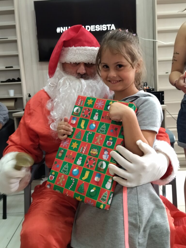 Imagem de uma menina sorridente segurando um presente embrulhado em papel colorido com estampas natalinas, como árvores de Natal, bonecos de neve e estrelas. Ao lado dela, está um Papai Noel sentado, vestindo a tradicional roupa vermelha com detalhes brancos e usando luvas brancas. Ele segura uma pequena sineta dourada em uma das mãos. 