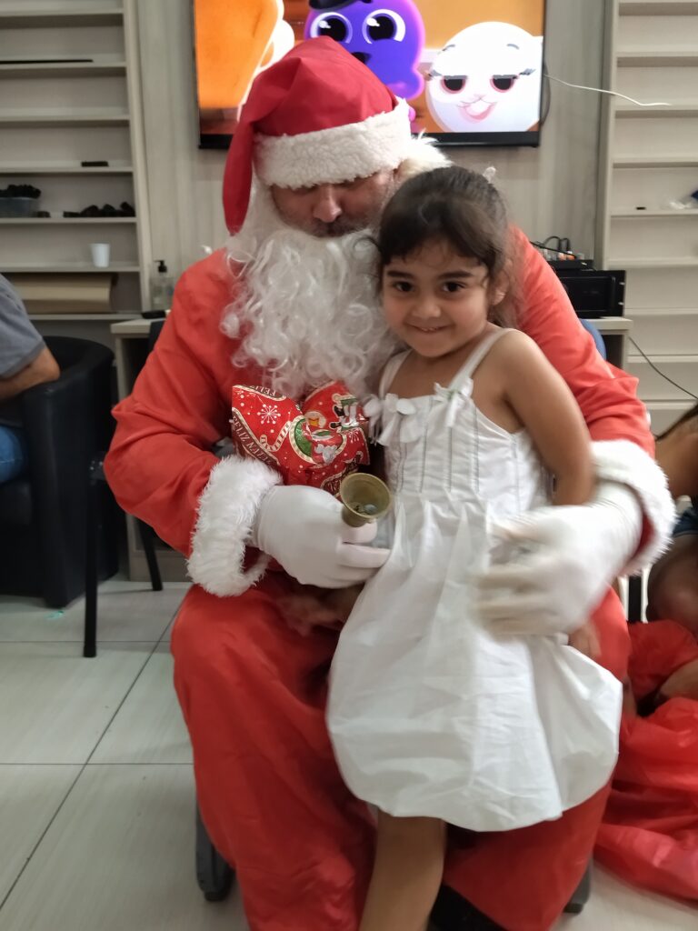 Imagem mostra uma cena de Natal, onde uma menina está sentada no colo do Papai Noel. A menina está sorrindo e vestindo um vestido branco. O Papai Noel está usando o tradicional traje vermelho com gorro e luvas brancas, e segura um pequeno sino dourado em uma das mãos.