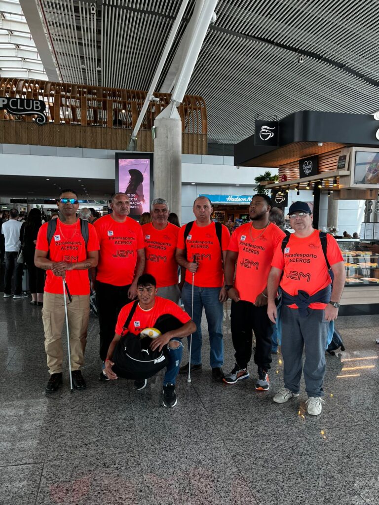 foto dos da equipe masculina de goalball. No aeroporto salgado filho, todos estão usando uma camiseta na cor laranja. Na frente da camiseta está escrito Paradesporto Acergs