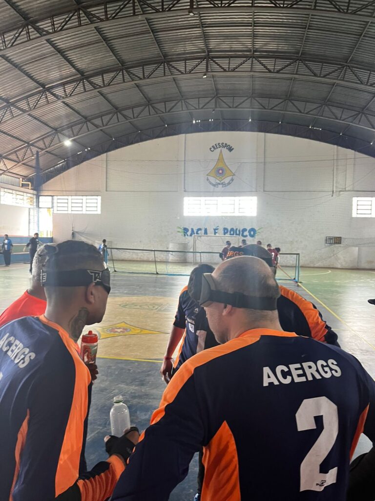 Quadra poliesportiva coberta, com teto de estrutura metálica e iluminação natural entrando pelas janelas altas. O foco principal está em um grupo de jogadores de goalball, vestindo uniformes azul escuro com detalhes laranja e a palavra "ACERGS" nas costas. Eles usam vendas pretas nos olhos, característica do esporte para cegos. Um dos atletas segura uma garrafa de água. Ao fundo, vê-se o gol do goalball e outros jogadores ou técnicos espalhados pela quadra. Na parede, há a logomarca da instituição "CIEESPOM" e a frase "RAÇA É POUCO" pintada em azul. O ambiente é de uma partida de goalball