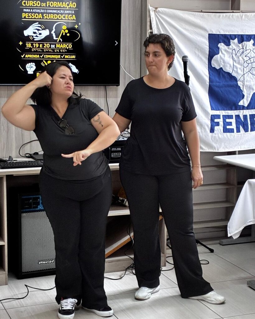 Duas mulheres, ambas vestidas de preto, estão em uma sala. Uma das mulheres faz gestos na Língua de Sinais  enquanto a  outra mulher observa ao lado e faz sinais de libra tátil na costas da primeira mulher que é uma pessoa surdocega . Atrás deles há uma tela com informações sobre um curso para comunicação com pessoas surdocegas e um banner da FENEIS.