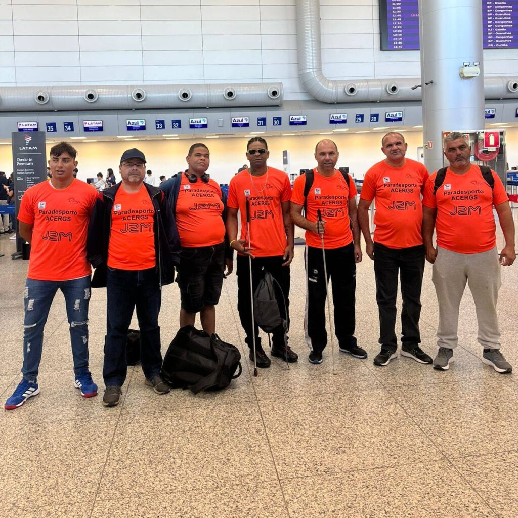 7pessoas com camisetas laranja escritas em azul “Paradesporto ACERGS” e “J2M” estão alinhadas em frente aos balcões de check-in da LATAM e Azul no aeroporto. Algumas seguram bengalas brancas e há mochilas no chão. Ao fundo, aparecem balcões numerados de 29 a 35, uma parede branca com dutos e um painel eletrônico de voos.