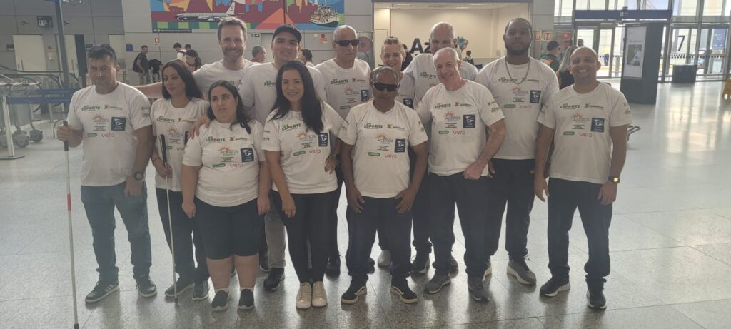 Grupo de 12 pessoas posando lado a lado em um saguão amplo de aeroporto.
Todas estão usando camisetas brancas iguais e calças ou bermudas escuras, parecendo um time ou delegação. Algumas pessoas têm bengalas brancas de mobilidade na mão, indicando que são pessoas cegas ou com baixa visão. Há homens e mulheres, com tons de pele variados do claro ao médio e escuro, idades aparentes de adultos jovens a mais velhos.
As camisetas trazem vários logotipos de projetos e patrocinadores na frente; em destaque, a palavra “ESPORTE” em verde, um sol estilizado com a palavra “ACERGS” abaixo e outros logotipos menores, incluindo “Vero” em vermelho. Várias pessoas estão com o braço apoiado no ombro de quem está ao lado, transmitindo união e clima de equipe.
Ao fundo, aparecem partes do aeroporto: piso claro, colunas, algumas pessoas passando, carrinhos de bagagem e um painel colorido grande na parede superior. A iluminação é forte e uniforme, vinda de janelas grandes ao fundo.