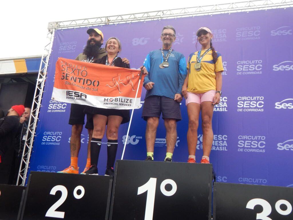 Quatro atletas sorridentes e medalhados posam no pódio de um painel azul escrito: “CIRCUITO SESC DE CORRIDAS”.
1º Lugar (Centro): Paulo Renato (homem branco, grisalho, de óculos, com camiseta longa azul e shorts escuro) e Janise (mulher branca, loira com cabelo preso, de camiseta, shorts e meias longas pretas). Janise segura uma faixa laranja e branca com desenhos de corredores e os textos: “PROJETO SEXTO SENTIDO Rio Grande do Sul”, “IESA Vamos juntos.” e “MOBILIZE FINANCIAL SERVICES”.
2º Lugar (Esquerda): Homem pardo de barba e bigode longos, usando boné amarelo, camiseta escura e tênis/meias laranja.
3º Lugar (Direita): Mulher morena clara de trança lateral, boné claro, camiseta amarela e azul, shorts rosa e tênis laranja.