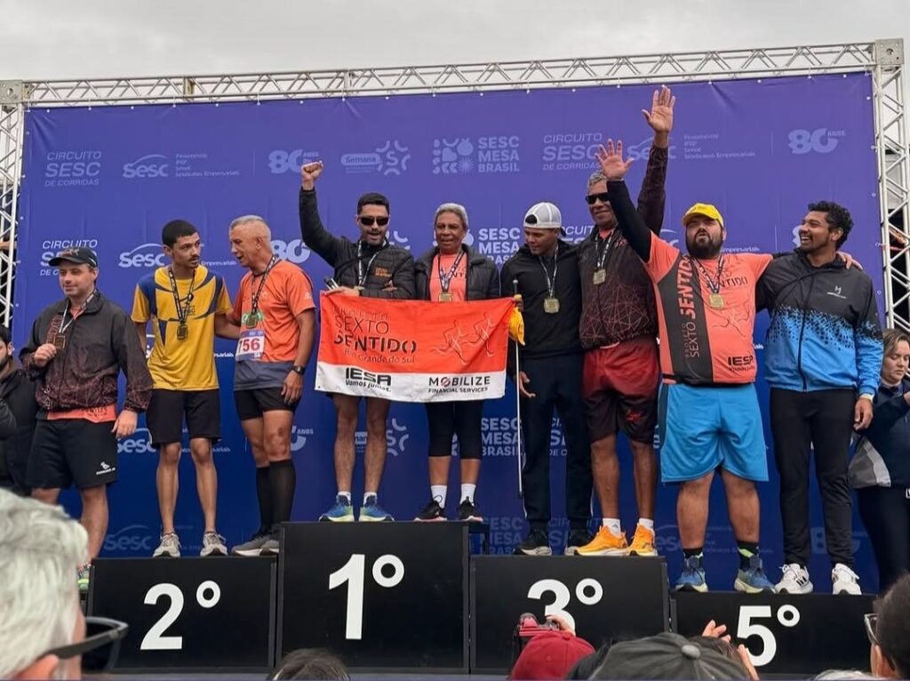Grupo de corredores sobre um pódio de premiação.
Ao centro está o bloco do 1º lugar, mais alto, com três pessoas em cima segurando uma faixa laranja e branca. Na faixa está escrito “Sexto Sentido – Rio Grande do Sul” e, abaixo, os logotipos “IESA” e “MOBILIZE PARAMAR SERVICES” com outros textos menores.
Os três do topo vestem roupas esportivas laranja, preta e cinza, todos com medalhas douradas penduradas no pescoço. O homem no meio ergue um dos braços em sinal de comemoração.
À esquerda, sobre o bloco marcado “2º”, há duas pessoas com medalhas, uma de camiseta amarela e azul e outra de camiseta laranja e número de peito 156.
À direita, sobre o bloco “3º”, estão duas pessoas com medalhas, uma de boné branco e roupa preta e outra com moletom escuro e bermuda vermelha.
Mais à direita, sobre o bloco “5º”, há dois homens: um com camiseta laranja, bermuda azul e boné amarelo, e outro com jaqueta azul e preta.
Ao fundo há um painel azul com os logotipos “SESC”, “Circuito Sesc de Corridas” e “SESC Mesa Brasil” repetidos.