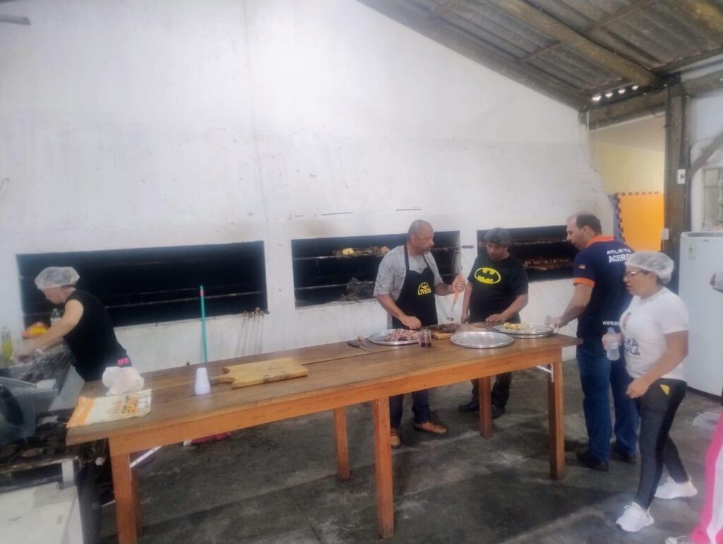 Cozinha ampla com um grande balcão de madeira no centro.
Ao fundo há uma longa churrasqueira embutida na parede branca, com várias aberturas retangulares escuras, e dentro delas alguns espetos com carnes assando.