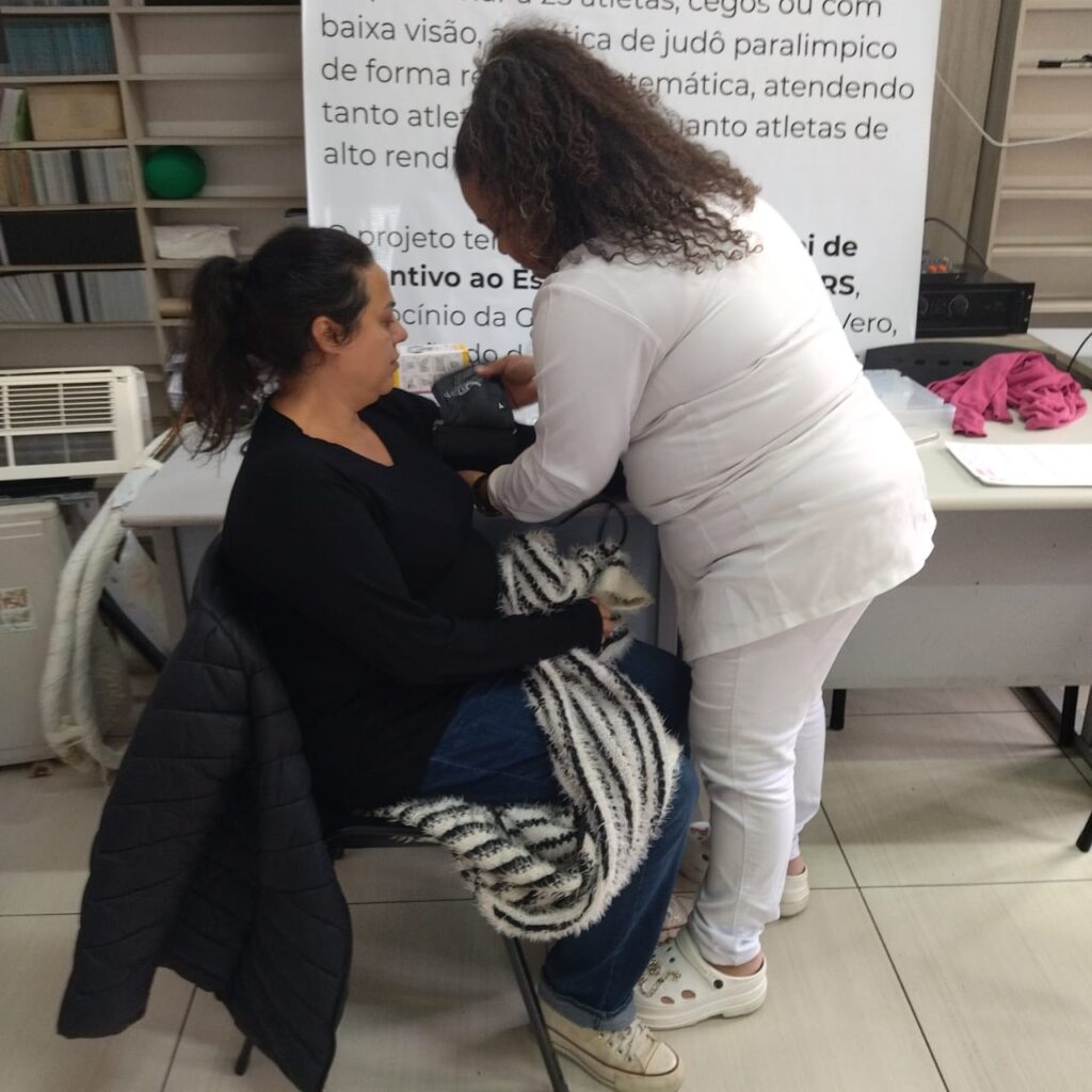 Duas mulheres estão em um ambiente interno de atendimento de saúde.
A mulher sentada, de pele clara e cabelo escuro preso, usa blusa preta e calça jeans, com uma manta listrada preto e branco no colo.
A outra mulher, de pele negra, cabelo cacheado e roupa branca de profissional de saúde, está em pé medindo a pressão da mulher sentada com um manguito no braço esquerdo dela.