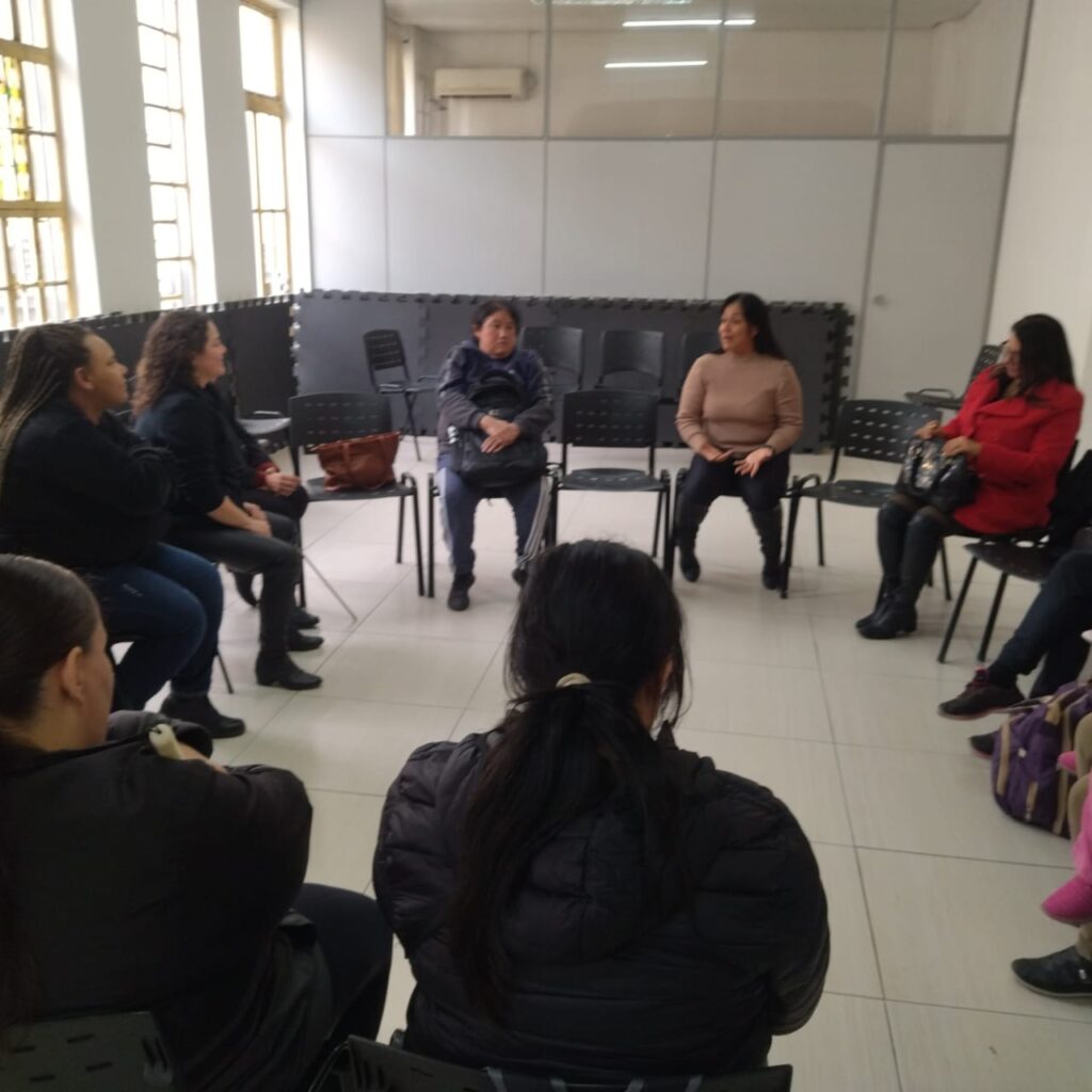 Salão amplo com várias mesas redondas ocupadas por pessoas sentadas em cadeiras plásticas brancas.
As mesas têm toalhas principalmente vermelhas e azuis, com pratos e copos descartáveis.
Há muitas pessoas adultas, homens e mulheres de idades variadas, algumas conversando em pé e outras sentadas comendo ou bebendo.