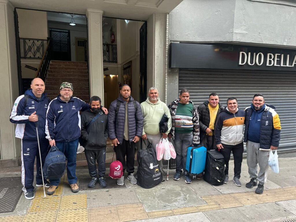 Grupo de dez homens adultos em pé em uma calçada em frente a um prédio.
Todos estão vestidos com roupas de frio, como jaquetas, moletons e calças compridas.
Quase todos carregam bagagens: mochilas nas costas, bolsas esportivas grandes e uma mala de rodinhas azul clara.
Do lado esquerdo, dois homens usam agasalhos esportivos azul‑marinho com detalhes brancos; um deles segura uma bengala branca e leva uma mochila azul na mão.
Ao lado deles há um homem de moletom preto com capuz e outro usando jaqueta acolchoada cinza e calça preta.
Mais ao centro está um homem de moletom verde‑claro segurando duas sacolas plásticas transparentes com itens dentro.
Perto dele há um homem com moletom colorido com estampa geométrica, calça escura e uma bengala.
À direita aparecem três homens sorrindo, usando jaquetas em tons de amarelo, branco, azul‑marinho e preto; um deles está junto da mala azul de rodinhas e outro segura uma sacola plástica branca.
Ao fundo está a entrada de um prédio com escadaria interna e corrimãos pretos de metal; ao lado direito há uma fachada com porta de metal fechada e o letreiro “DUO BELLA” em letras claras sobre placa preta.