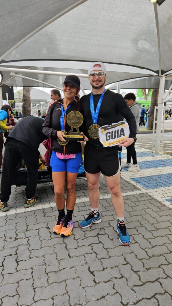 Duas pessoas posam após uma corrida segurando troféus.
À esquerda, uma mulher de pele clara bronzeada, boné e jaqueta pretos, shorts azuis e tênis de cores diferentes (um laranja e um amarelo). Ela segura um troféu dourado grande em forma de medalha sobre pedestal e usa outra medalha azul no pescoço.
À direita, um homem de pele clara, boné branco para trás, óculos escuros, camisa e shorts pretos, tênis esportivos azuis. Ele usa uma medalha dourada igual e segura uma placa branca e amarela com a palavra “GUIA” em letras grandes pretas. Estão em área de chegada de prova de rua, com outras pessoas ao fundo.