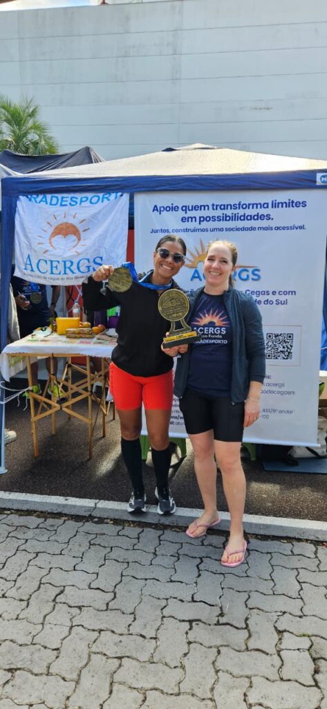 Duas mulheres sorrindo em frente a uma tenda da ACERGS em um evento ao ar livre.
A mulher à esquerda tem pele morena, cabelo preso para trás e usa óculos escuros. Ela segura uma grande medalha dourada pendurada em uma fita azul e, com a outra mão, um troféu dourado com base escura e um círculo grande na parte de cima.
A mulher à direita tem pele clara, cabelo loiro preso para trás e veste camiseta azul‑marinho com o logotipo da ACERGS e a palavra “PARADESPORTO”. Ela está encostada de leve na outra mulher, em pose de comemoração.
Atrás delas há uma tenda com lona azul e branca. À esquerda, um banner com o símbolo de um sol alaranjado e o texto “ACERGS Associação de Cegos do Rio Grande do Sul”.
Ao centro e à direita, um grande painel com o texto em azul: “Apoie quem transforma limites em possibilidades. Juntos, construímos uma sociedade mais acessível.” Abaixo, novamente o logotipo com o sol laranja e a palavra “ACERGS” em azul, seguido de outras linhas de texto menores sobre a associação e um código QR.
