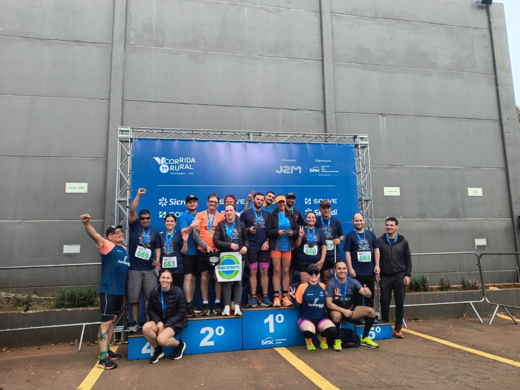 Um grupo grande de corredores posando em um pódio de prova de rua em frente a um painel azul.  
As pessoas estão em três níveis de pódio com indicações “4º”, “2º” e “1º” na base, todas usando medalhas azuis penduradas no pescoço e números de peito presos às camisetas.  
A maioria veste camiseta azul de corrida, alguns com detalhes laranja, shorts ou leggings esportivas e tênis coloridos; há homens e mulheres de diferentes tons de pele, em clima de comemoração, alguns com braço erguido, outros fazendo sinal de positivo ou abraçados.  
Uma pessoa no centro segura um círculo verde e branco com o texto “FUNESPORTE”.  
No painel azul atrás aparecem vários logotipos e textos, incluindo “7ª CORRIDA RURAL”, “Fertisystem – J2M”, além de marcas como Sicredi e Sesc.