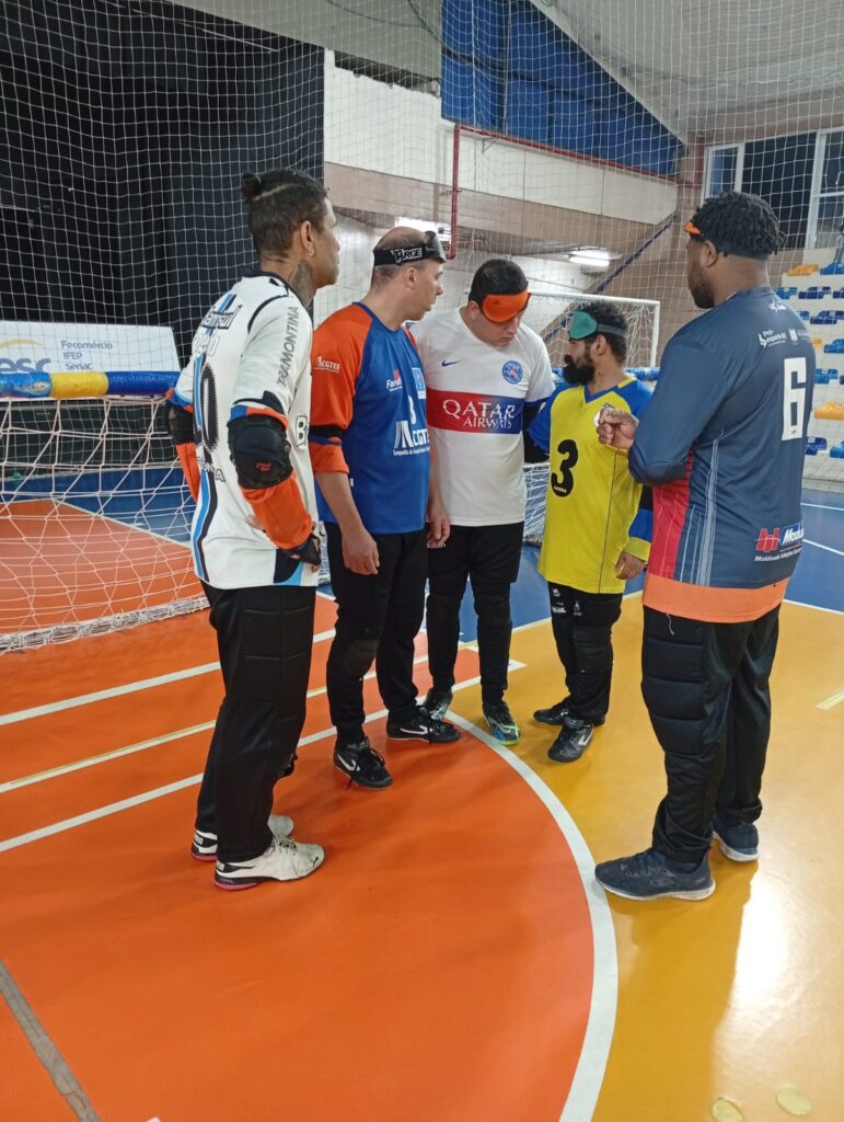 Cinco atletas de goalball estão em uma quadra coberta de piso laranja e amarelo. Eles usam uniformes esportivos, calças acolchoadas e vendas nos olhos. Estão posicionados diante de uma grande rede, aparentando conversar ou se preparar para o jogo.