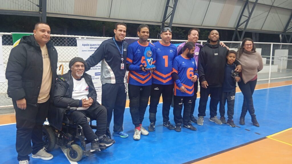 Premiação da equipe masculina de goalball da ACERGS em uma quadra coberta. Ao centro, três jogadores com uniforme azul e laranja, usando vendas, posam segurando um troféu. Ao redor deles estão organizadores do evento e, ao fundo, o técnico da equipe, todos sorridentes.