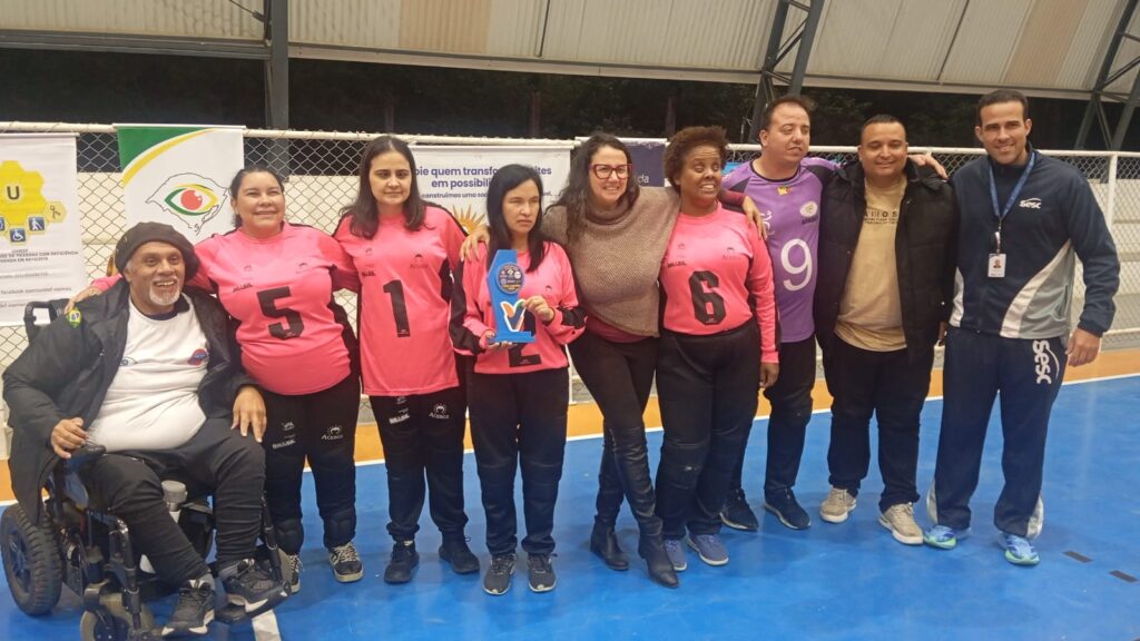 Equipe feminina de goalball da ACERGS vice-campeã, posando em quadra coberta. Da esquerda para a direita: um homem negro em cadeira de rodas; três atletas com uniforme rosa e preto, números 5, 1 e 2 (esta segurando o troféu azul); uma mulher de óculos abraçada ao grupo; outra jogadora com número 6; um atleta com uniforme roxo número 9; um homem de jaqueta preta; e o técnico da equipe, com agasalho azul do Sesc e crachá. Todos estão sorrindo, sobre o piso azul da quadra, com banners ao fundo.