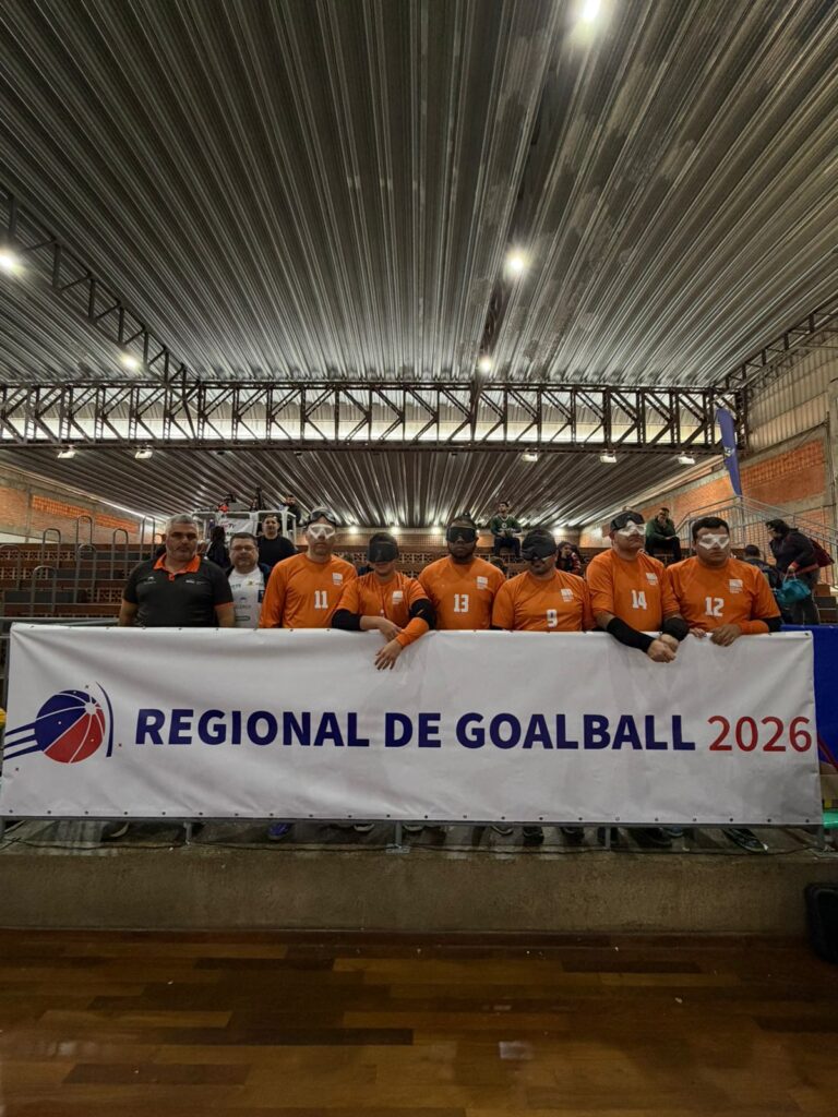 Banner branco grande de campeonato de goalball em um ginásio coberto.
À frente do banner, está uma equipe de jogadores de goalball usando camisas laranja com números brancos no peito e vendas ou óculos escuros esportivos.
Ao lado esquerdo dos jogadores há um homem de camisa polo preta com detalhes laranja, aparentando ser da comissão técnica.
Atrás do grupo principal há mais algumas pessoas em pé, com roupas escuras, em um nível um pouco mais alto, como em arquibancadas metálicas.
O banner na frente deles traz, à esquerda, um logotipo redondo com curvas em azul e vermelho e, ao lado, o texto em letras grandes azuis: “REGIONAL DE GOALBALL” e, em seguida, em vermelho: “2026”.