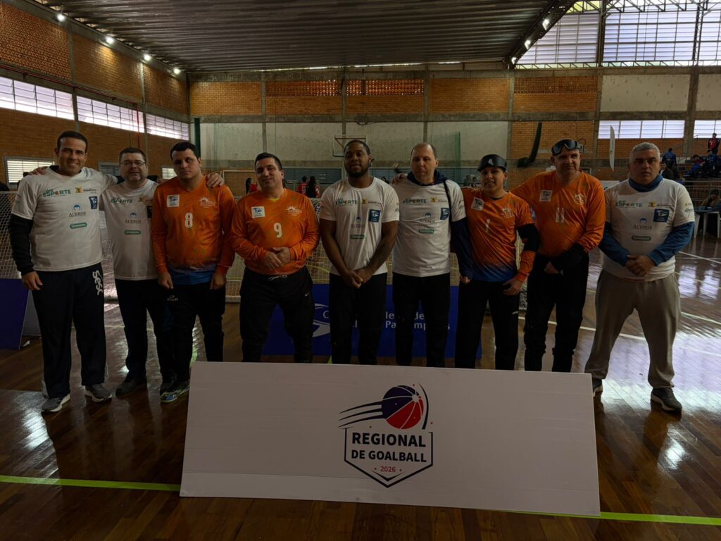 Oito pessoas em pé em uma quadra esportiva. Atrás delas há um painel branco escrito Regional de Goalball 2026. Quatro atletas usam uniforme laranja com números 8, 9, 1 e 11 e vendas nos olhos. Quatro pessoas usam camisetas brancas com logotipos e a palavra ACERGS. Ao fundo, público sentado em arquibancada dentro de um ginásio.