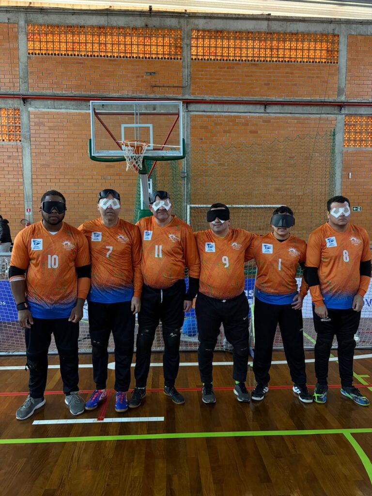 Seis atletas de goalball com deficiência visual, lado a lado, usando uniformes laranja e azul numerados e vendas nos olhos em ginásio.