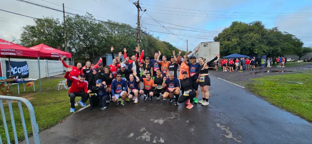 Grupo de cerca de vinte corredores de rua posa sorrindo e acenando em uma rua de asfalto úmido. Homens e mulheres de idades diversas vestem roupas esportivas e números de peito. À esquerda, há uma tenda vermelha da "Multifeira Restinga" e uma faixa escrita "Corre Restinga". Ao fundo, aparecem um caminhão baú branco, outras pessoas dispersas, árvores e céu nublado.