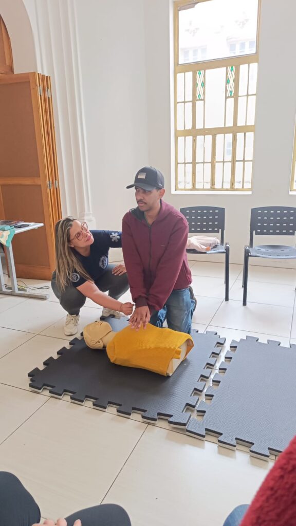 Sala ampla e clara com um treinamento de primeiro socorros em andamento.
No centro da imagem, Flávio de pele morena clara está ajoelhado sobre um tatame de EVA preto. Ele usa boné preto, jaqueta de moletom bordô com zíper e calça jeans azul. As mãos dele estão entrelaçadas e posicionadas no centro do tórax de um boneco de treinamento de reanimação cardiopulmonar, que tem cabeça e braços cor de pele clara e está coberto por um colete ou manta amarela.
À esquerda do Flávio, Jo de pele clara e cabelos loiros compridos, soltos, está agachada ao lado dele. Ela usa óculos de armação grossa, camiseta azul-marinho com um símbolo branco na manga e calça cinza, com tênis claros. Jo segura e ajusta o posicionamento das mãos do Flávio no peito do boneco, em atitude de orientação, enquanto o observa atentamente.
O tatame preto está montado em placas encaixadas sobre o piso de cerâmica clara.
