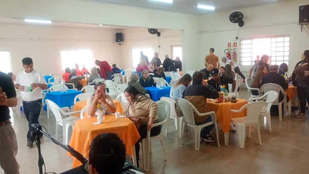 Salão amplo, com várias mesas cobertas por toalhas azuis e laranjas e cadeiras brancas. Há muitas pessoas conversando e confraternizando. Ao fundo, há janelas, ventiladores e extintores de incêndio. No primeiro plano, aparece um teclado musical com microfone, sugerindo que há música no ambiente. O clima é descontraído e de festa.


